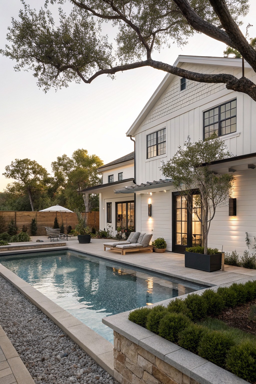 White two-story farmhouse with black window trim and a stone retaining wall bordering a long rectangular pool.