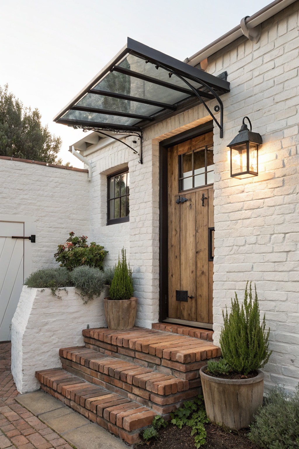 White brick house entrance with wooden door, glass awning, brick steps, and planters