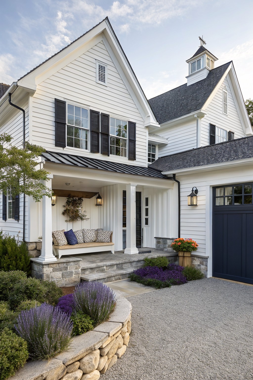 A white two-story farmhouse exterior with a covered front porch, white columns, a wooden bench with cushions, black shutters, and stone steps leading to a gravel driveway.