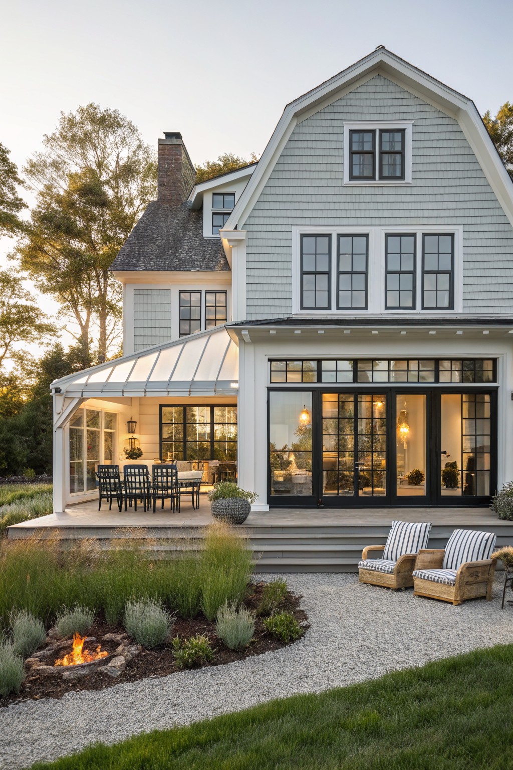 A light gray shingled house with a large wooden deck, black-framed windows and doors, a stone fire pit in a planted bed, and two striped armchairs on a gravel surface beside the deck.