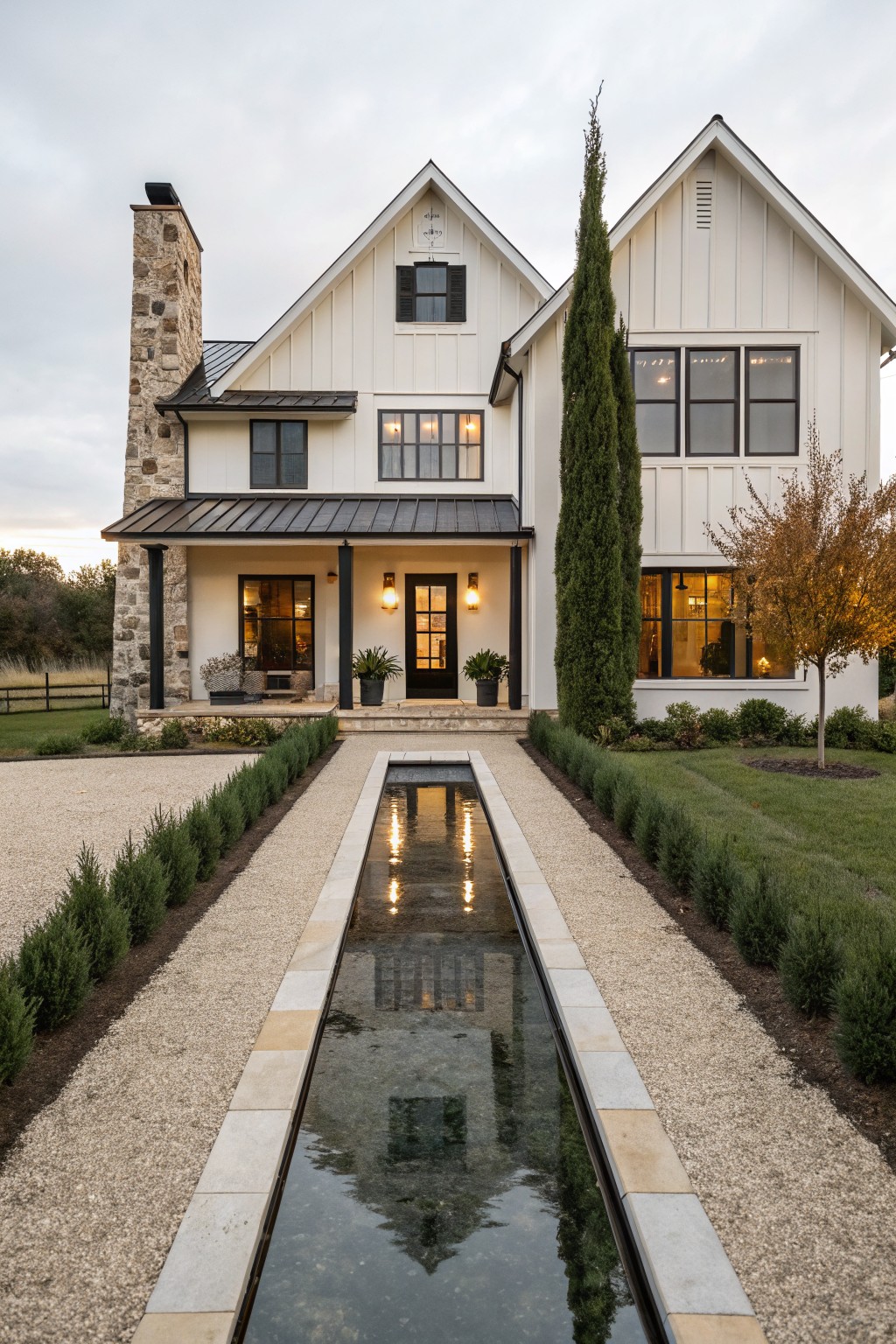 A modern farmhouse exterior with white vertical siding, a stone chimney, and a long rectangular reflecting pool centered between gravel paths leading to the front door.
