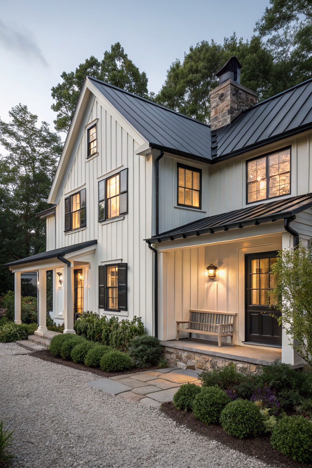 White two-story farmhouse with black roof, lit windows, and stone chimney at dusk.