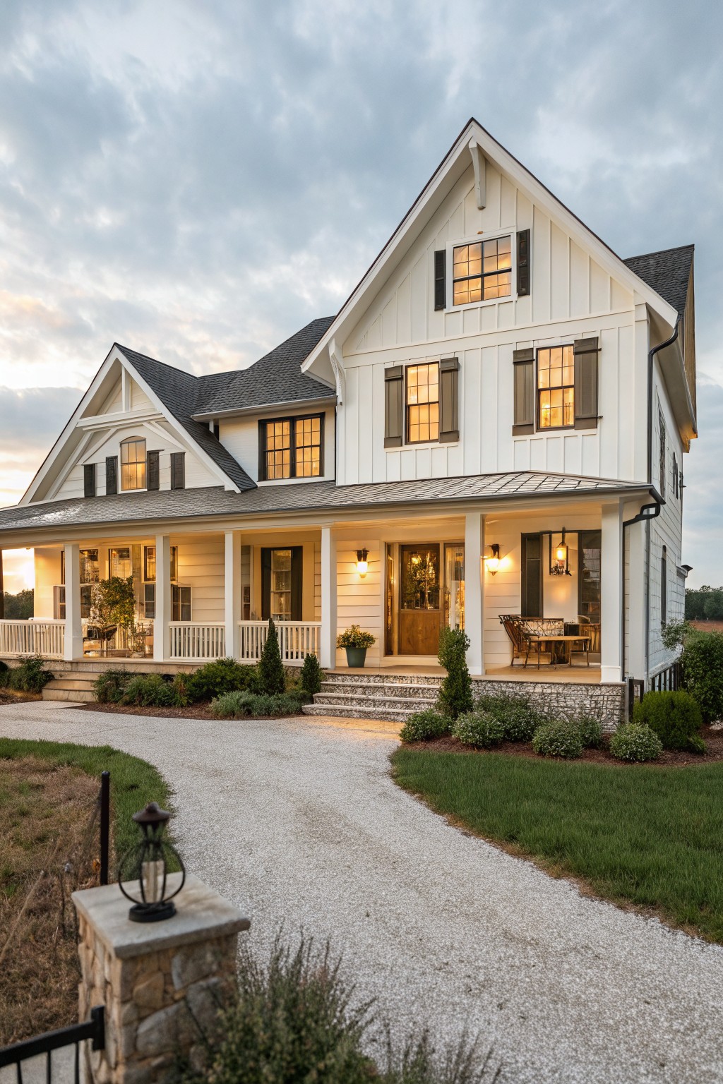 A two-story white farmhouse with dark wood shutters, a wooden front door, and a stone porch base viewed from a gravel driveway at dusk.