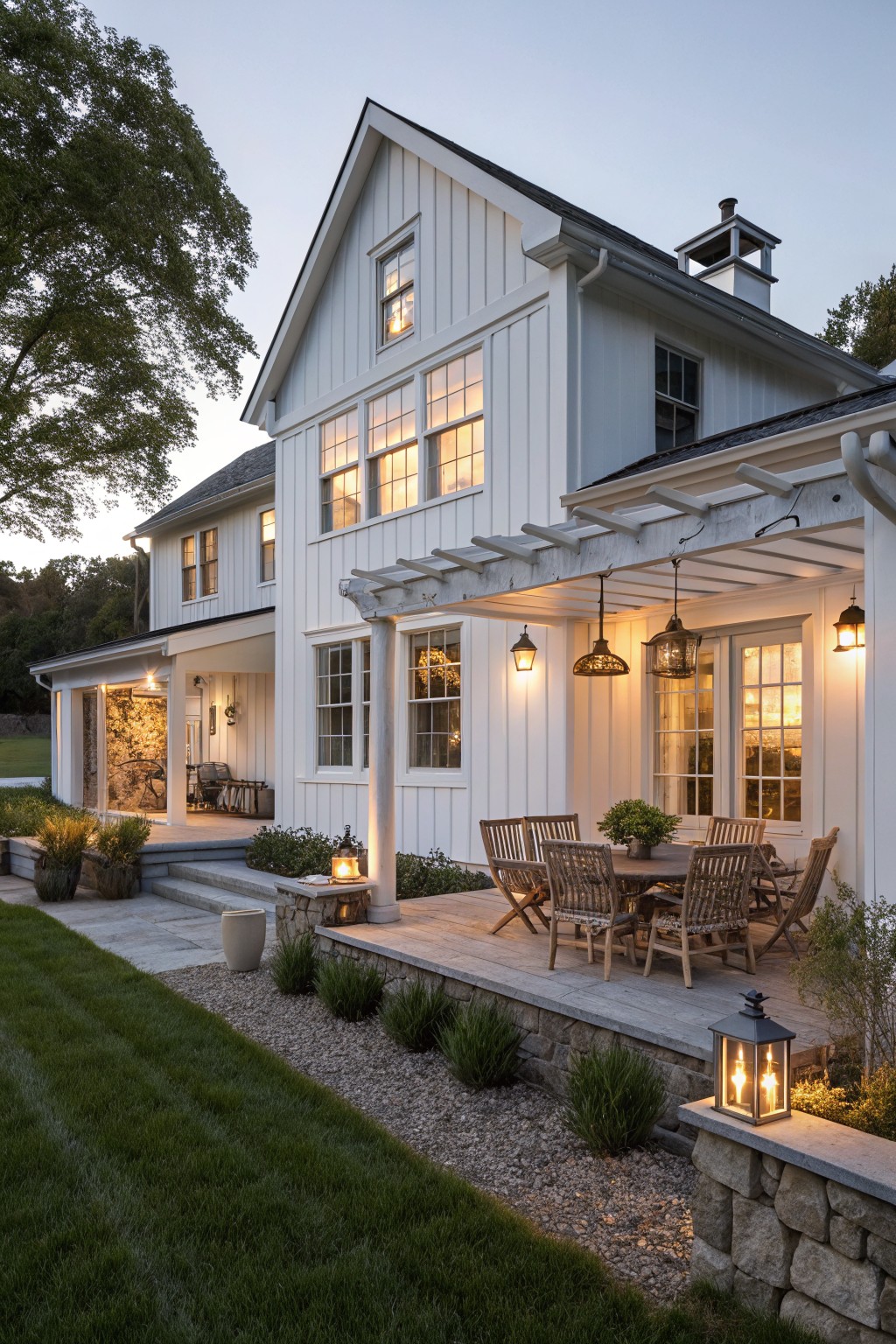 A white two-story farmhouse with a stone foundation has a wooden patio under a white pergola, set with a dining table and chairs, and illuminated by hanging lanterns at dusk.