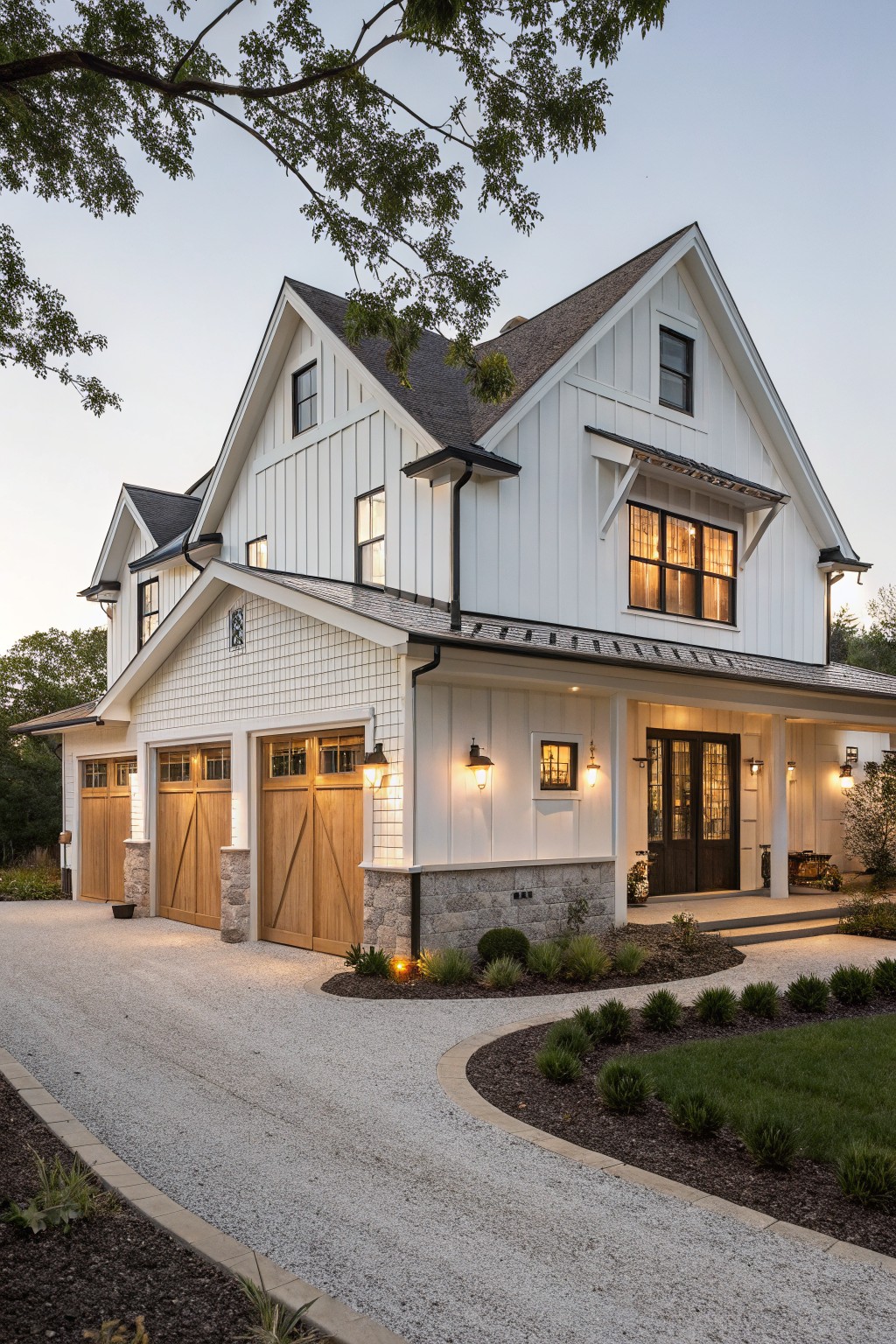 A white two-story farmhouse with multiple gables, board and batten siding, wooden garage doors, a stone foundation, and a covered porch entrance.