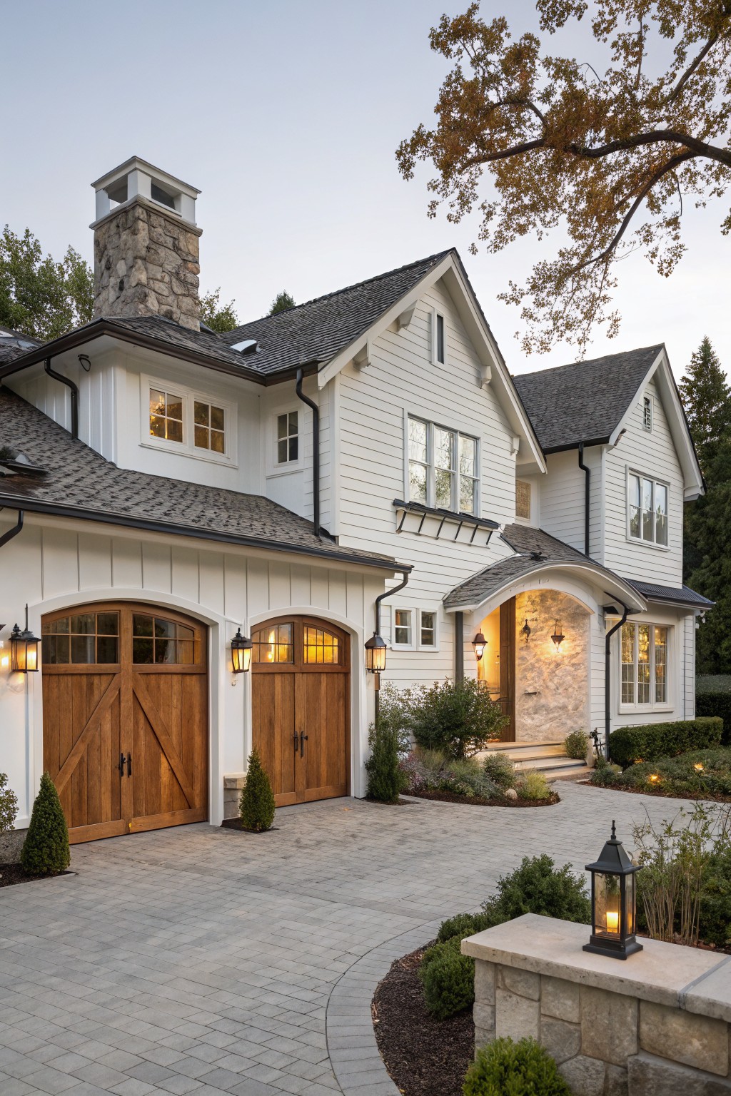 Elegant white two-story home with wooden garage doors, stone chimney, and lit lanterns.