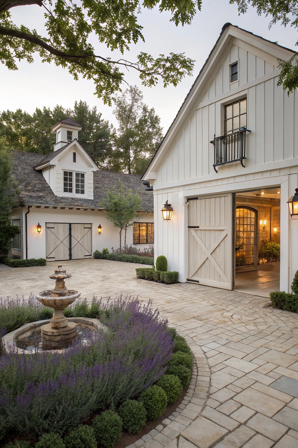 A white farmhouse exterior with a paved courtyard, stone fountain, and multiple carriage-style garage doors set into the facade.