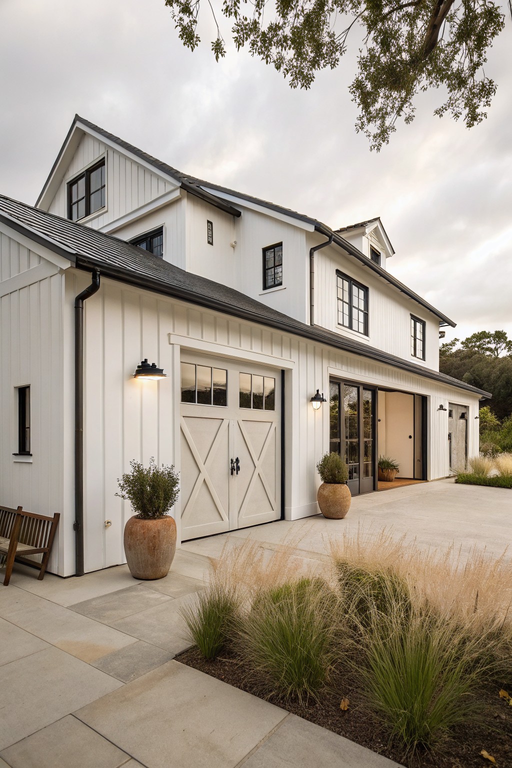 A white two-story farmhouse with vertical siding, black trim, and two integrated garage doors including a barn-style double door with X panels.