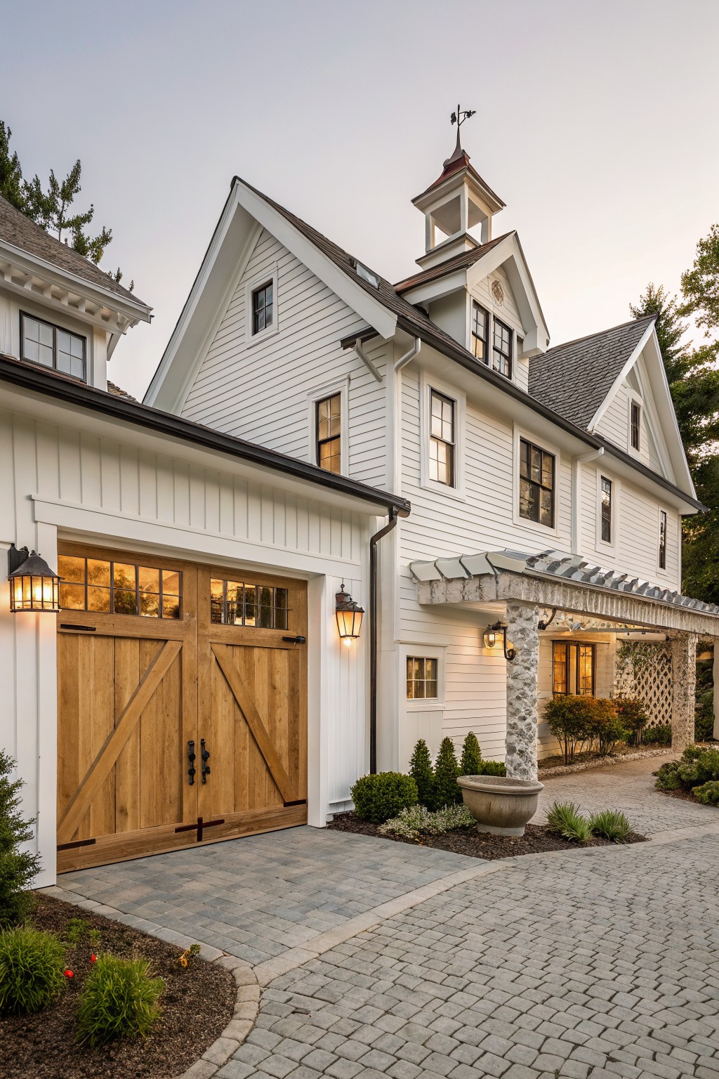 White house with wooden barn doors, lanterns, and cobblestone driveway at dusk.