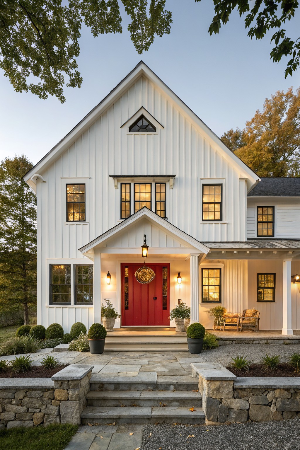 A white vertical-siding farmhouse with a red front door, black window frames, a small covered porch, and stone steps leading up from a gravel driveway.
