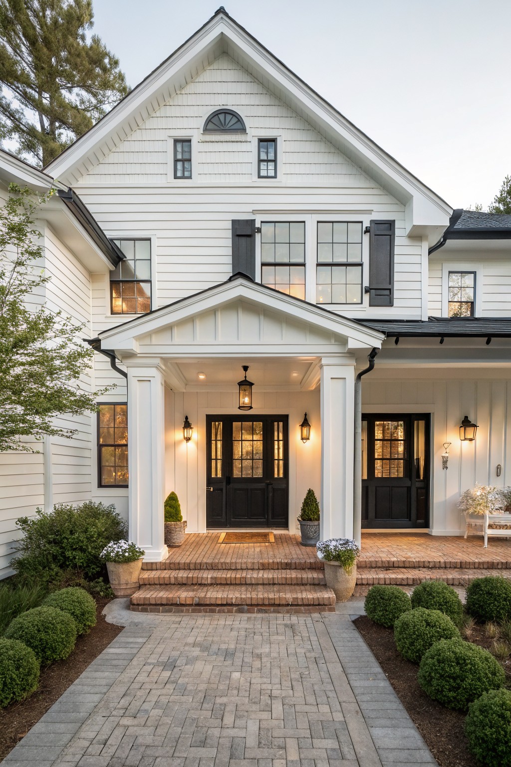 Front view of a white two-story farmhouse with black double doors, black shutters, a covered porch with brick steps, and potted plants along the walkway.