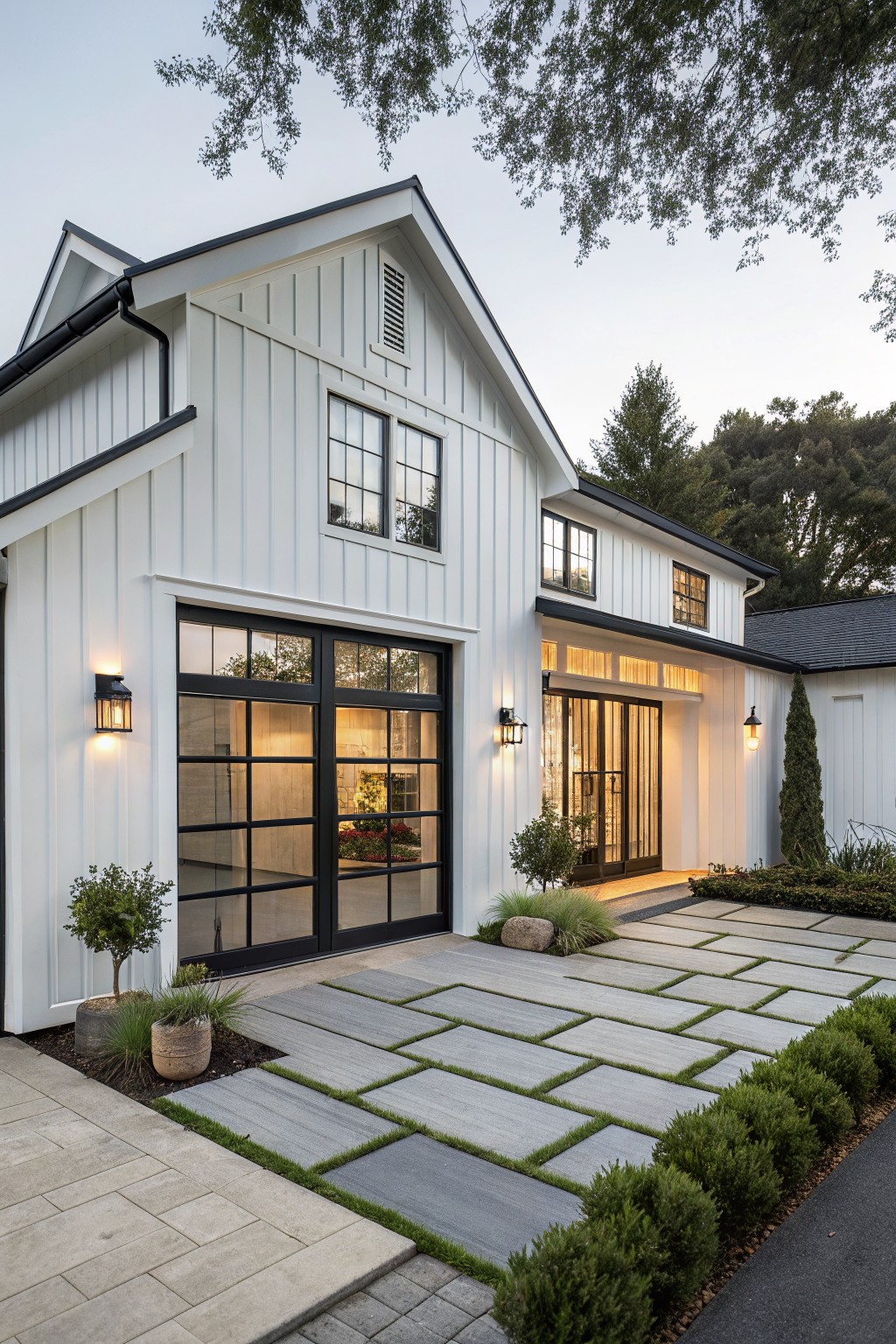 White modern farmhouse with black trim, large glass doors, stone pavers, and evening lights.