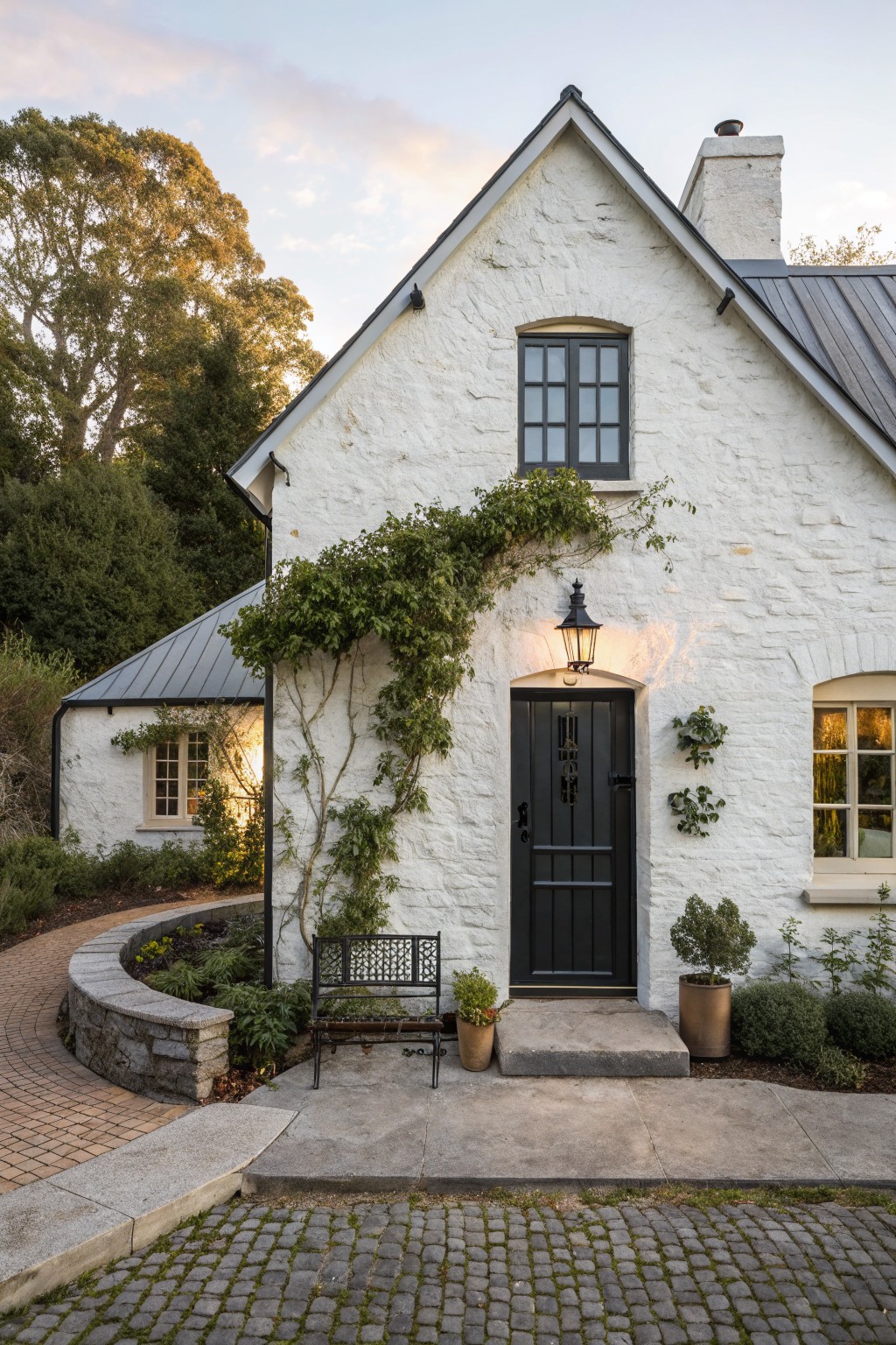 A white stucco farmhouse with a black front door, black window frames, and green climbing vines draping over the entrance, plus a black bench and potted plants on the steps.