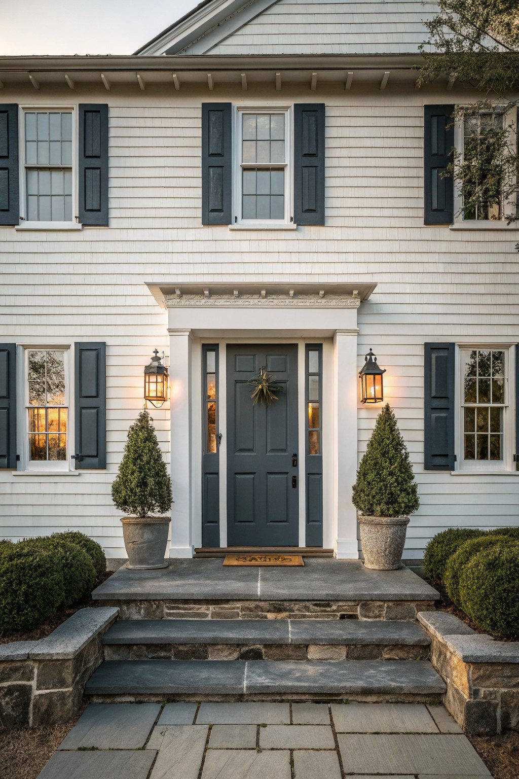 White house entrance with dark gray door, shutters, potted trees, and lanterns.