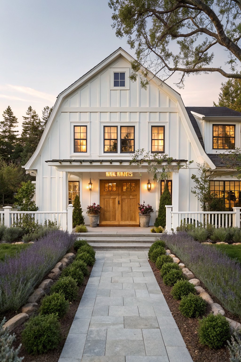 A white barn-style farmhouse exterior with a centered wooden double door, lit windows, and a stone walkway bordered by lavender and shrubs.