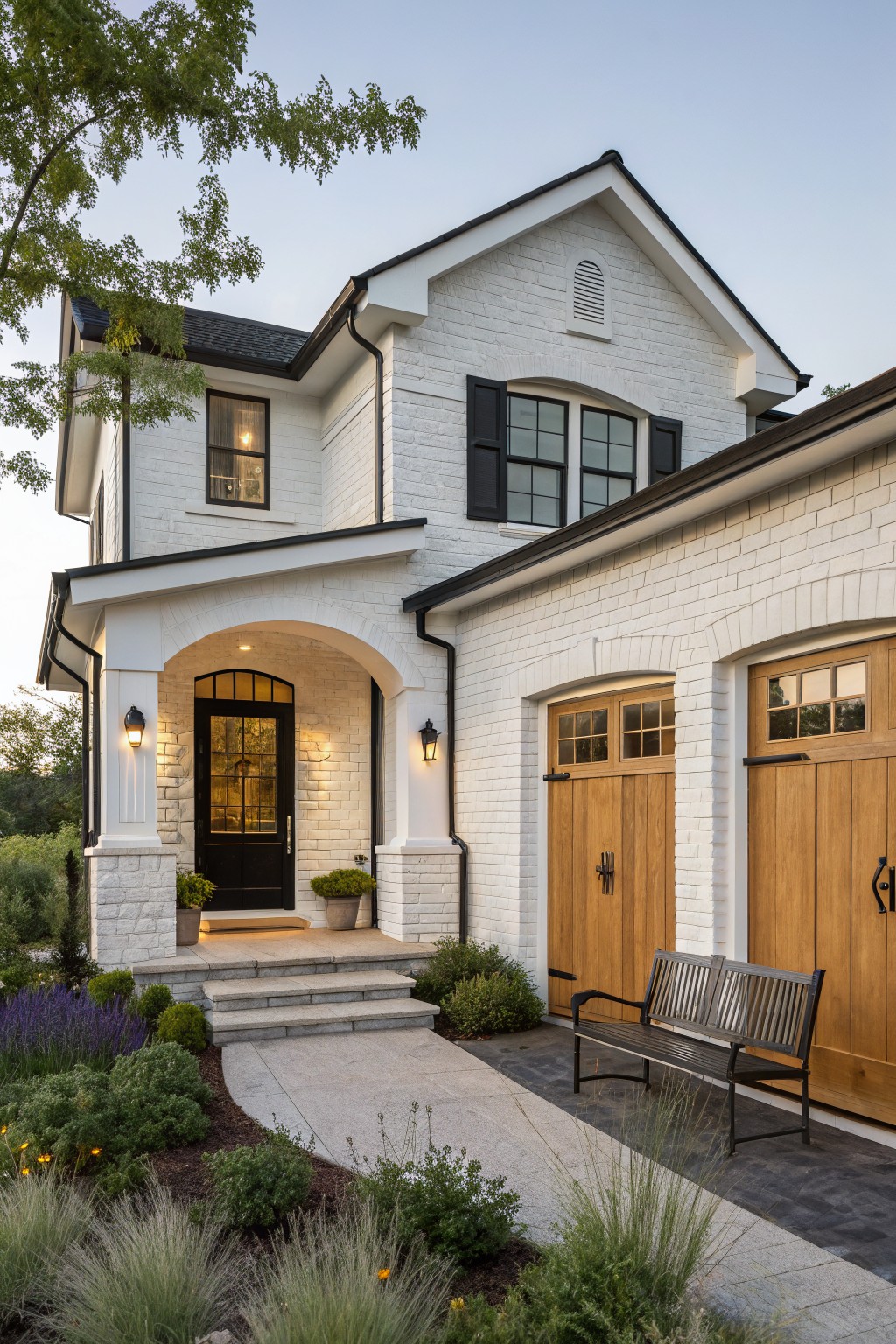 White brick house with arched entry, wooden garage doors, bench, and garden at dusk