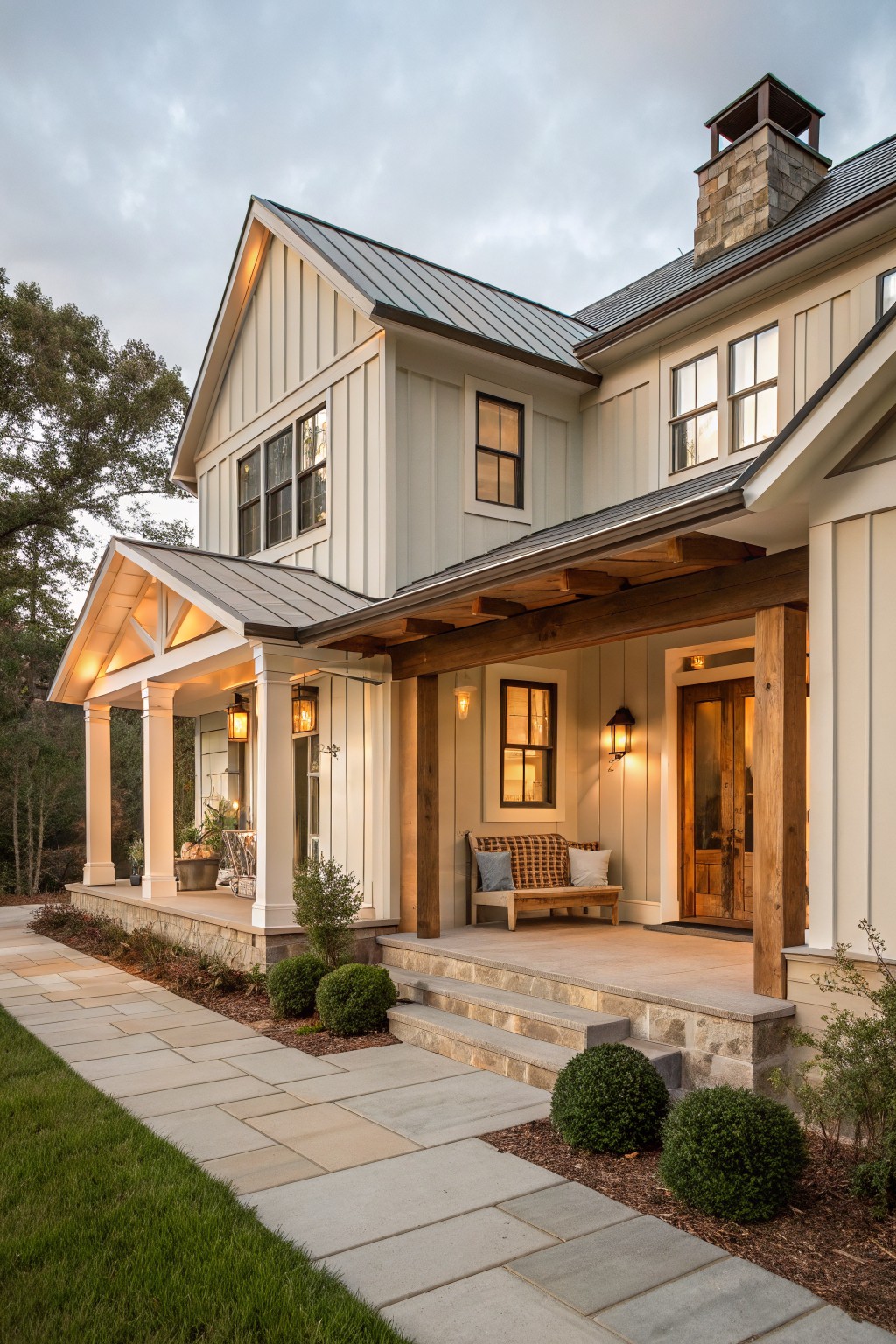 A two-story house exterior with light siding, a dark metal roof, wooden porch beams and posts, a stone chimney, and steps leading to a covered entry.