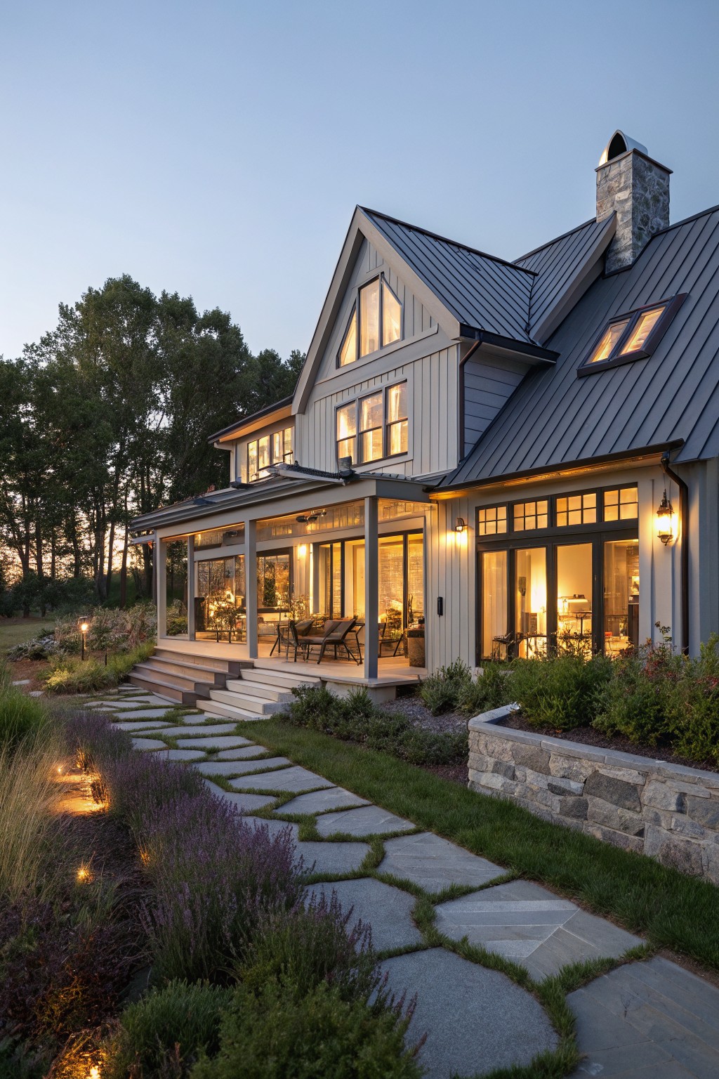A two-story coastal farmhouse with dark metal roofing, light siding, a stone chimney, and a stone pathway leading through landscaped garden beds at dusk.