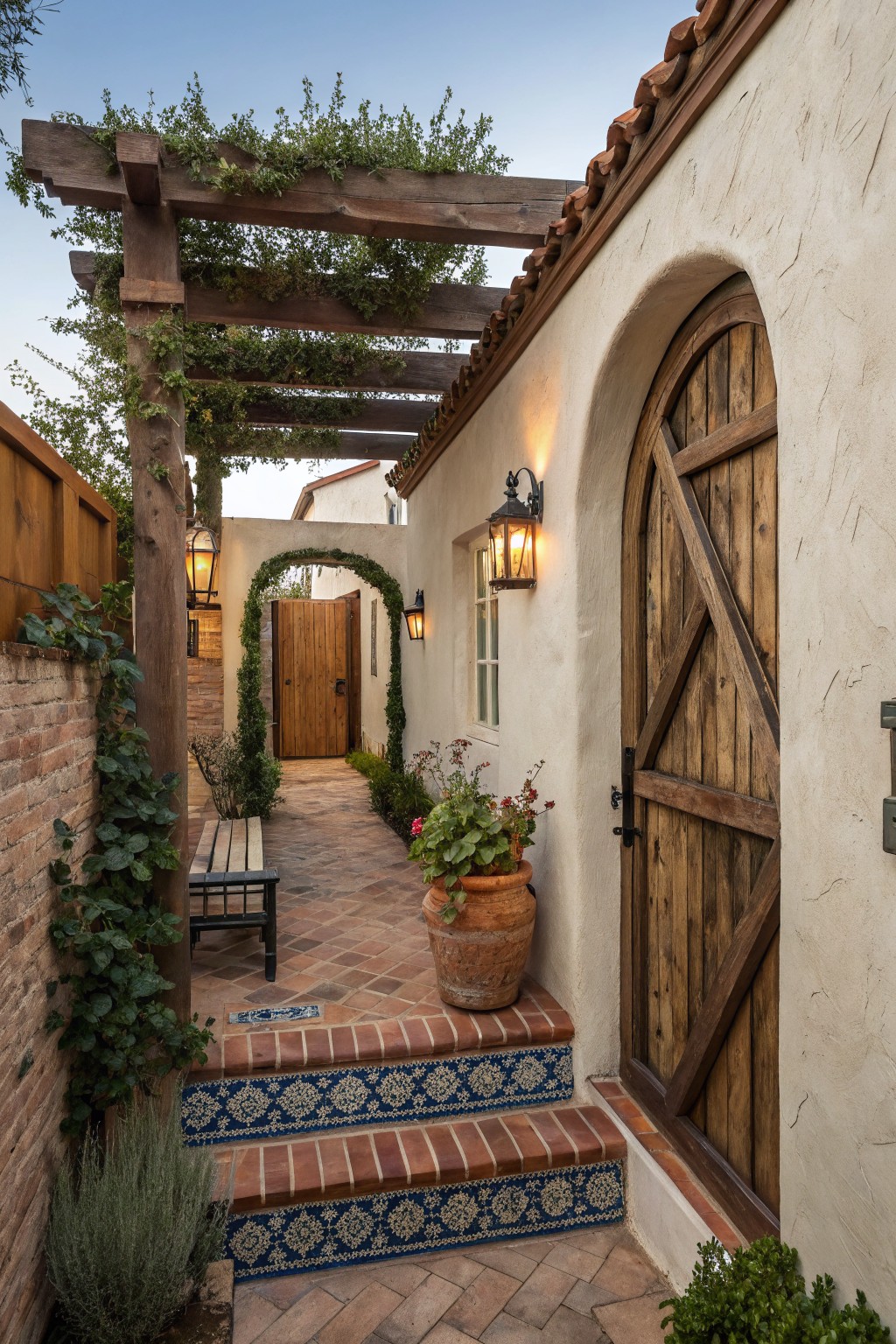 A narrow brick path with patterned tile steps leads to a large wooden door set in an arched opening on a stucco wall, with a wooden pergola overhead and potted plants nearby.