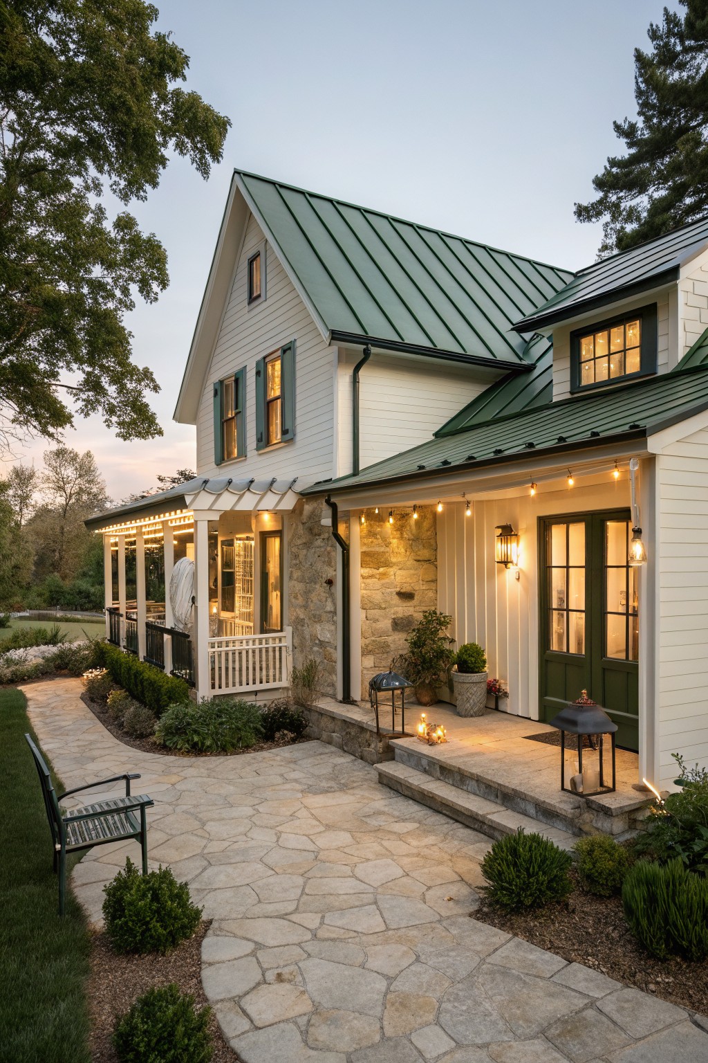 A compact white farmhouse with green metal roofing, green shutters, a stone base, and a front porch strung with lights.
