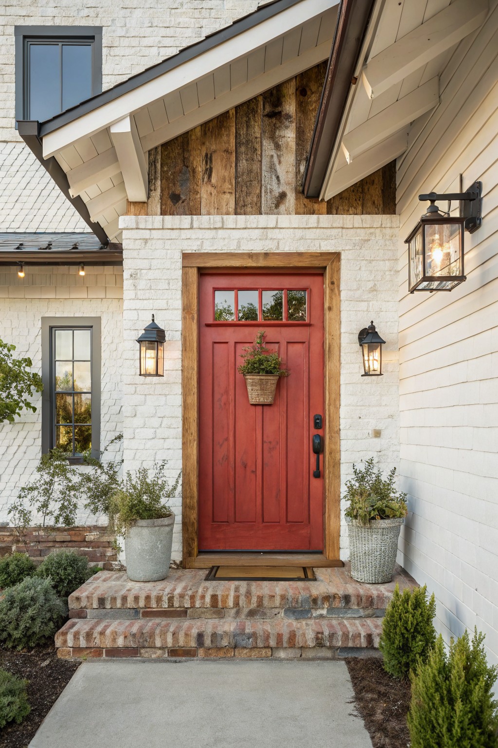 Front view of a white brick house entrance featuring a red door with a natural wood frame, two lit wall lanterns, and potted plants on brick steps.