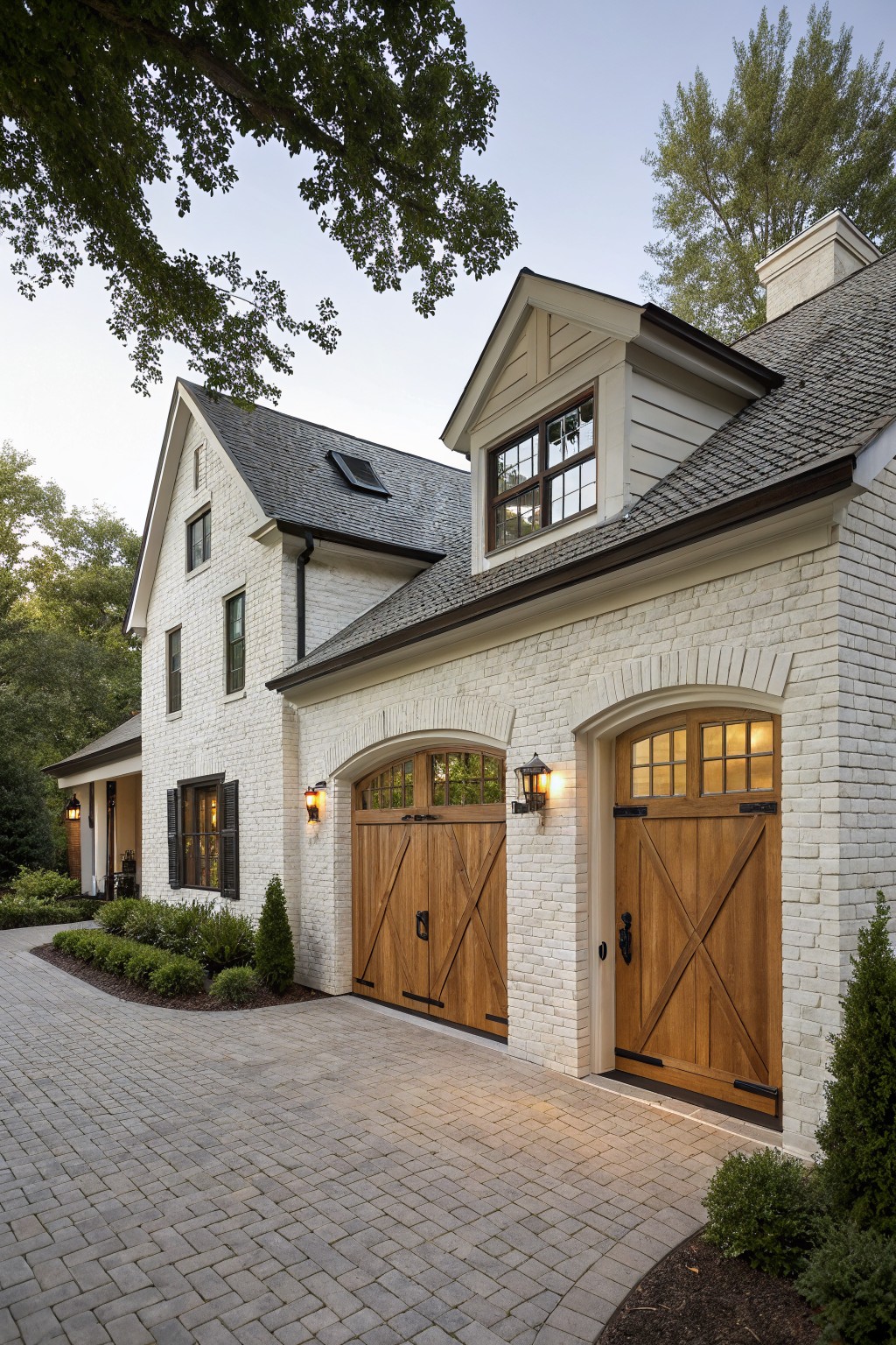 A white brick farmhouse exterior featuring two wooden garage doors with arched tops and cross bracing, set along a brick driveway with landscaping and trees nearby.