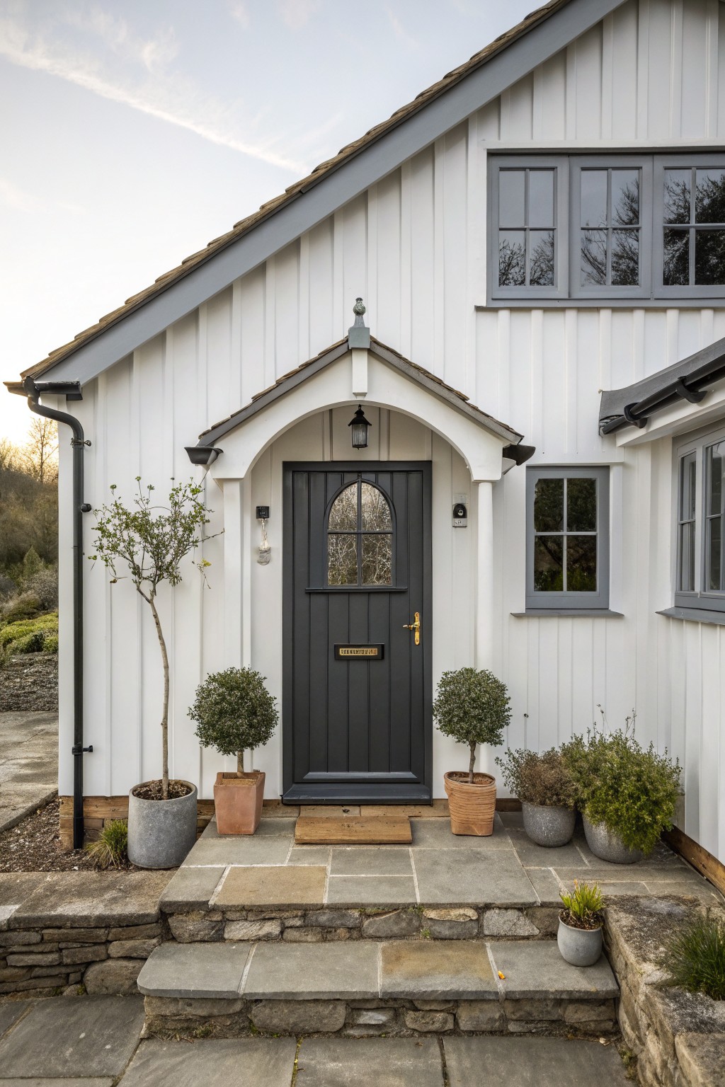 Front view of a white vertical-siding cottage with a dark gray door set under a small arched entry, stone steps, and potted plants on either side.