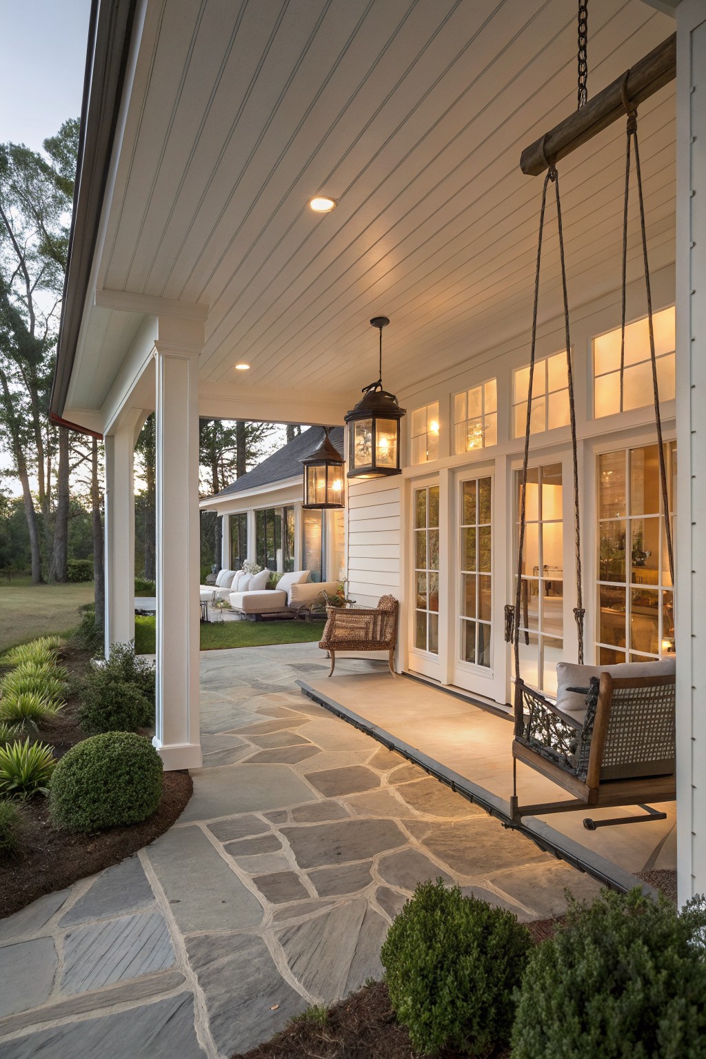 A covered farmhouse porch with a wooden hanging swing, stone paver floor, white siding and doors, and two dark metal lanterns.