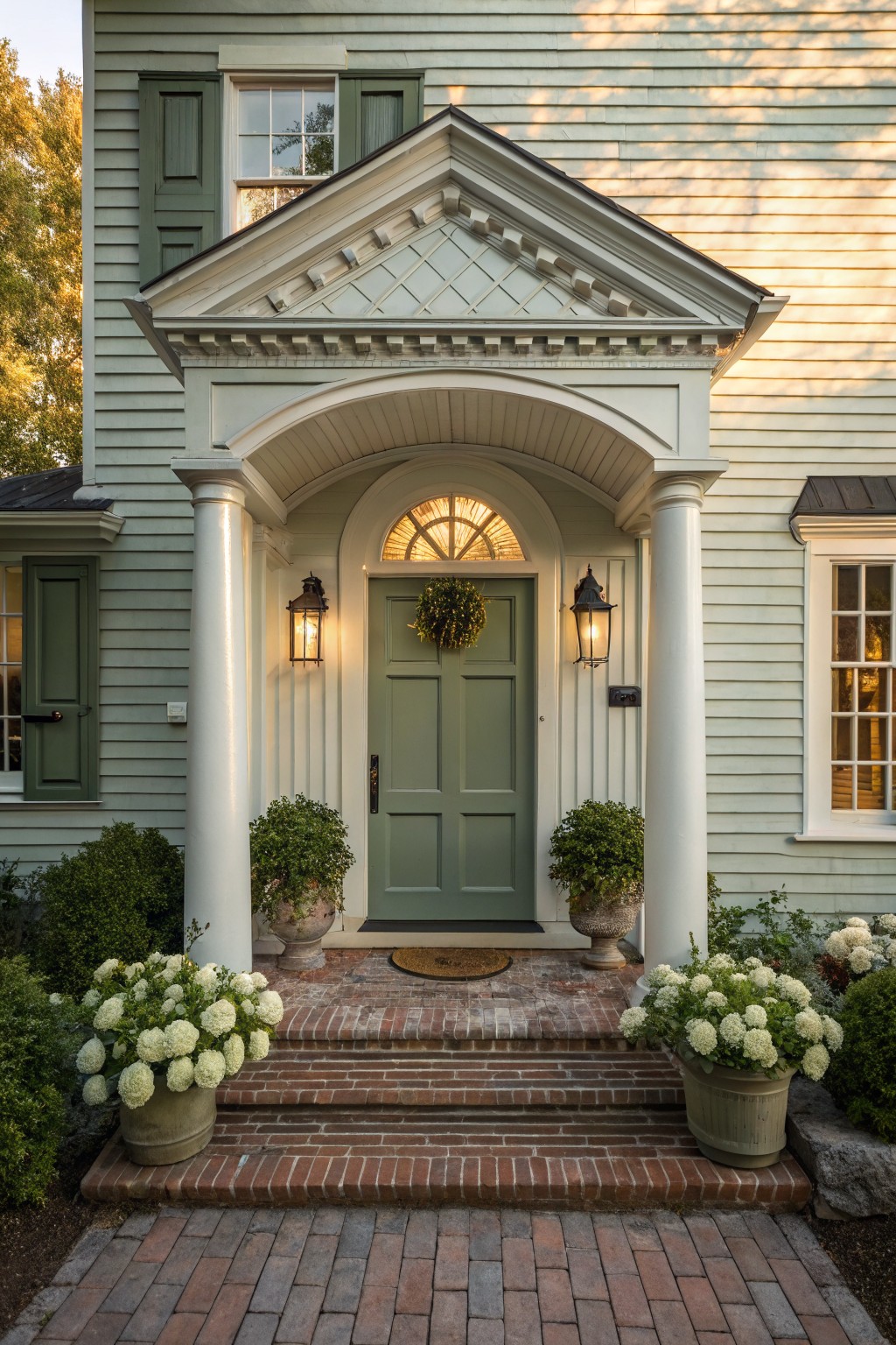 A light green house exterior with an arched white portico entrance supported by two columns, a green front door with a wreath, wall lanterns, and brick steps with potted plants on either side.
