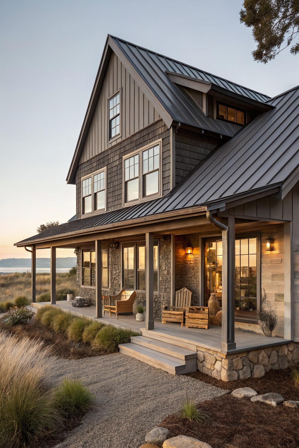 A two-story house with dark gray vertical siding, a dark metal roof, stone accents on the porch supports, and a covered front porch with steps leading to a gravel path.