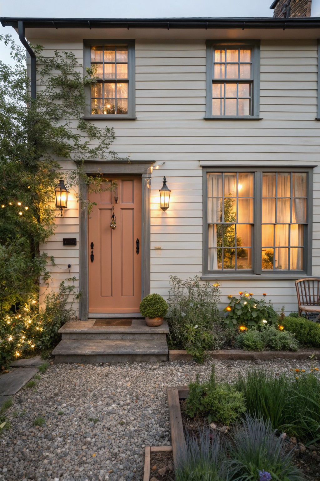 A two-story house with light siding features a coral front door, gray window trim, stone steps, and a gravel front yard with plants and string lights.