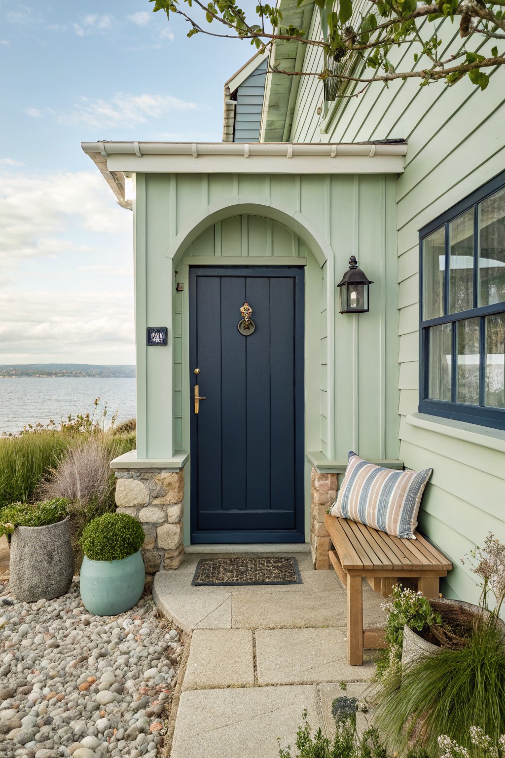 A light green shingled house exterior with a dark blue front door set under an arched entry, stone accents at the base, and a wooden bench to the right.