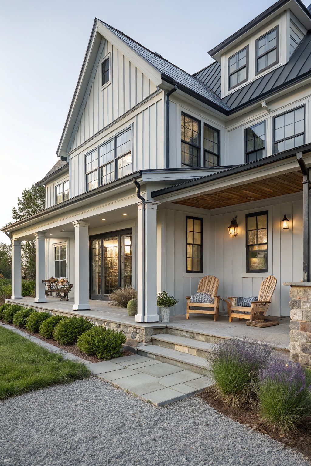 A two-story white house with vertical siding, a dark metal roof, multiple windows, and a covered front porch with wooden chairs and stone steps.