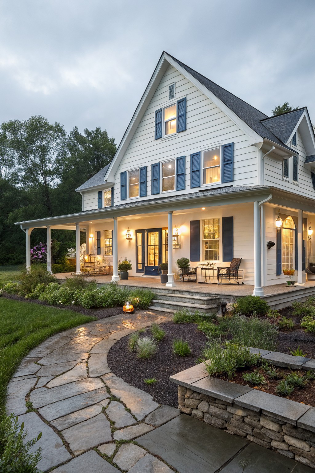 A white two-story farmhouse with blue shutters features a large wrap-around porch with multiple seating areas, a curved stone pathway, and garden beds in the foreground.