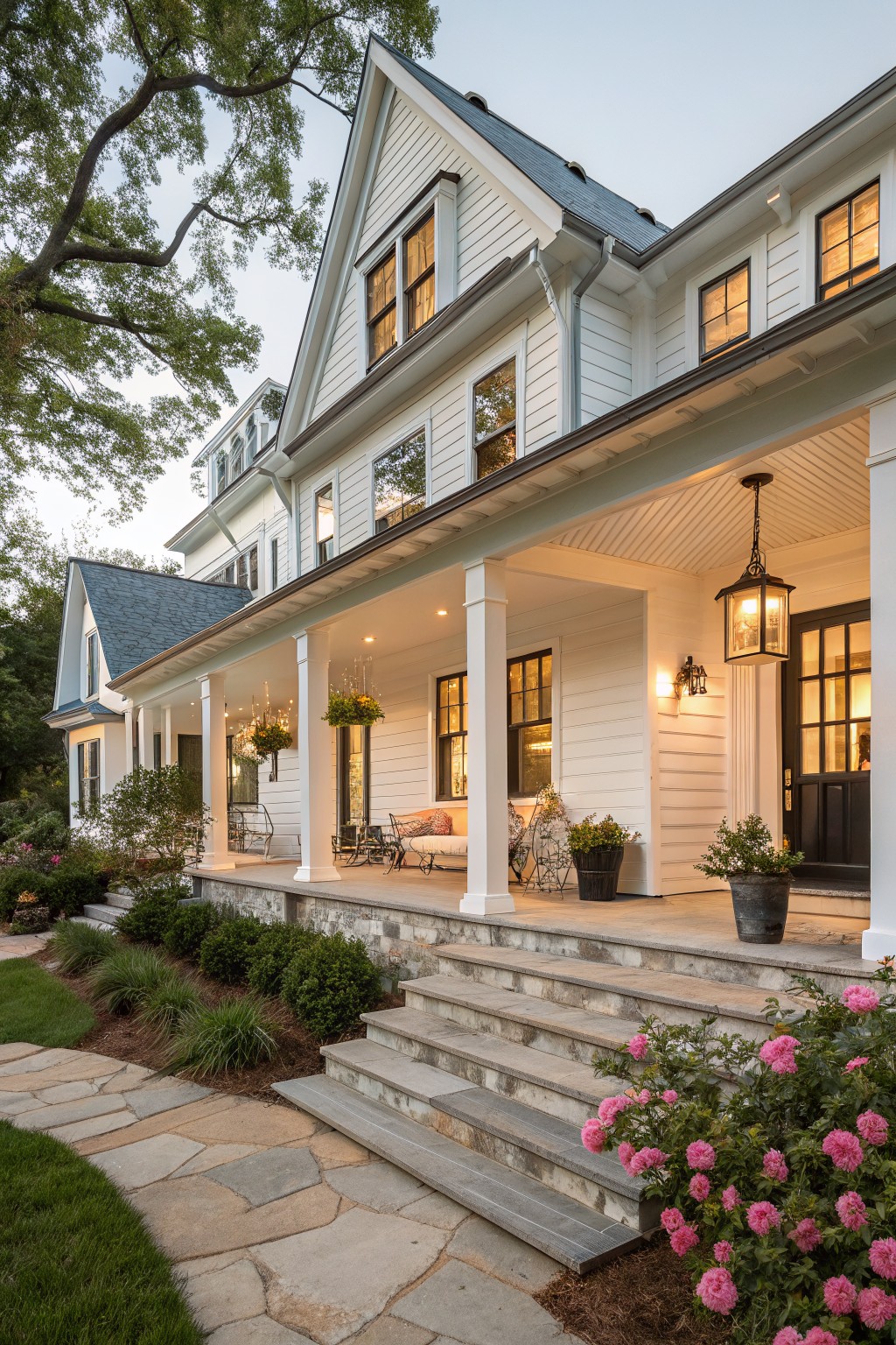 A white two-story farmhouse with a wrap-around porch, white columns, stone steps, and pink flowering shrubs in the foreground.