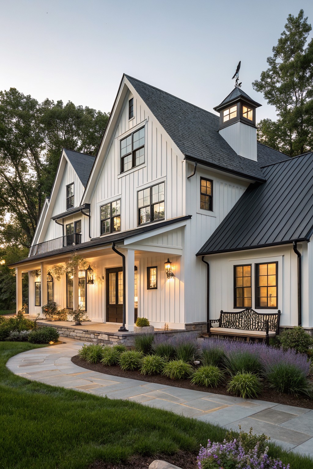 A white two-story farmhouse with black trim, black metal roof, and a small cupola, viewed from a curved stone pathway with landscaping in the foreground.