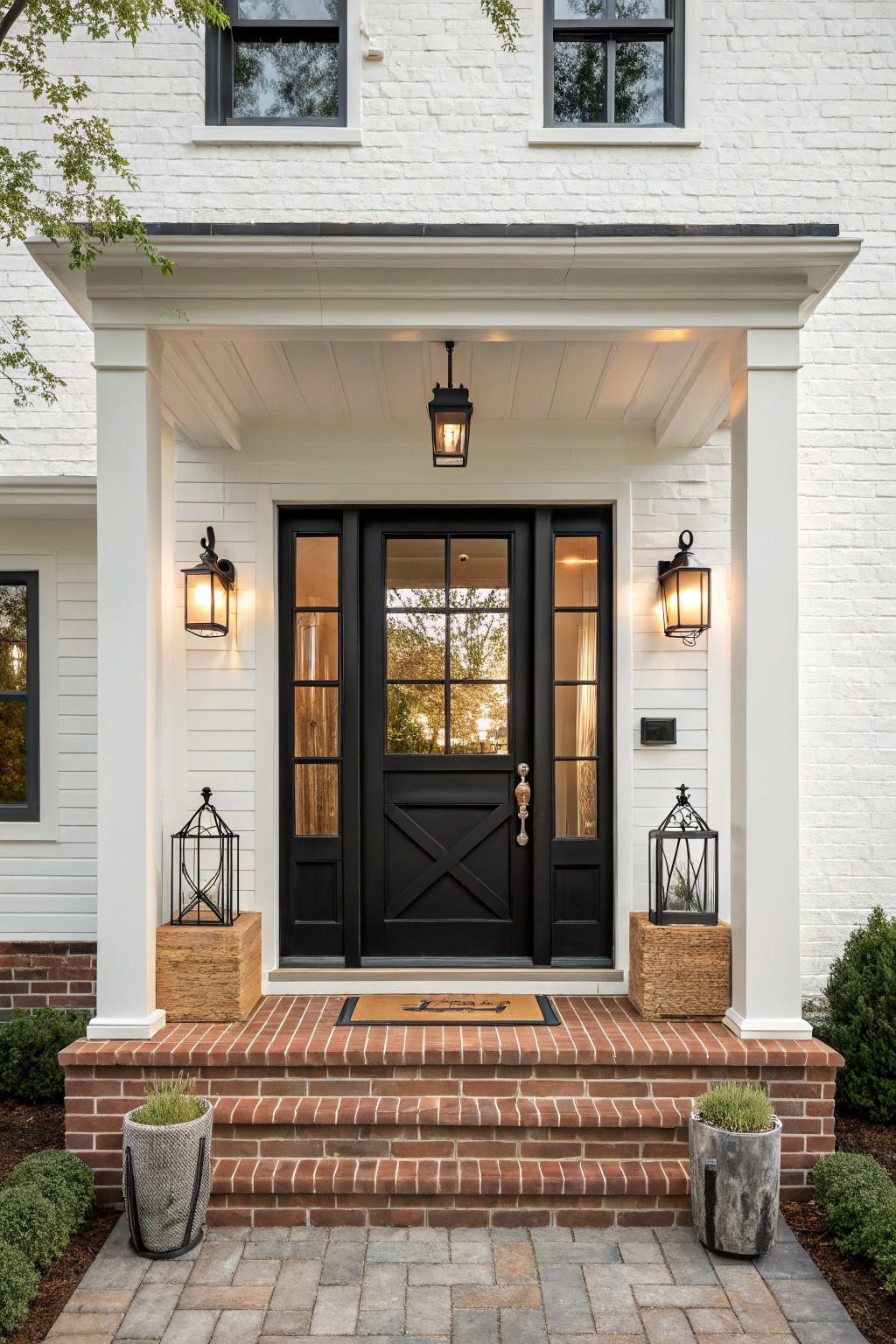 Front view of a white brick house with a black door, black window trim, and black lanterns on a covered porch with brick steps.