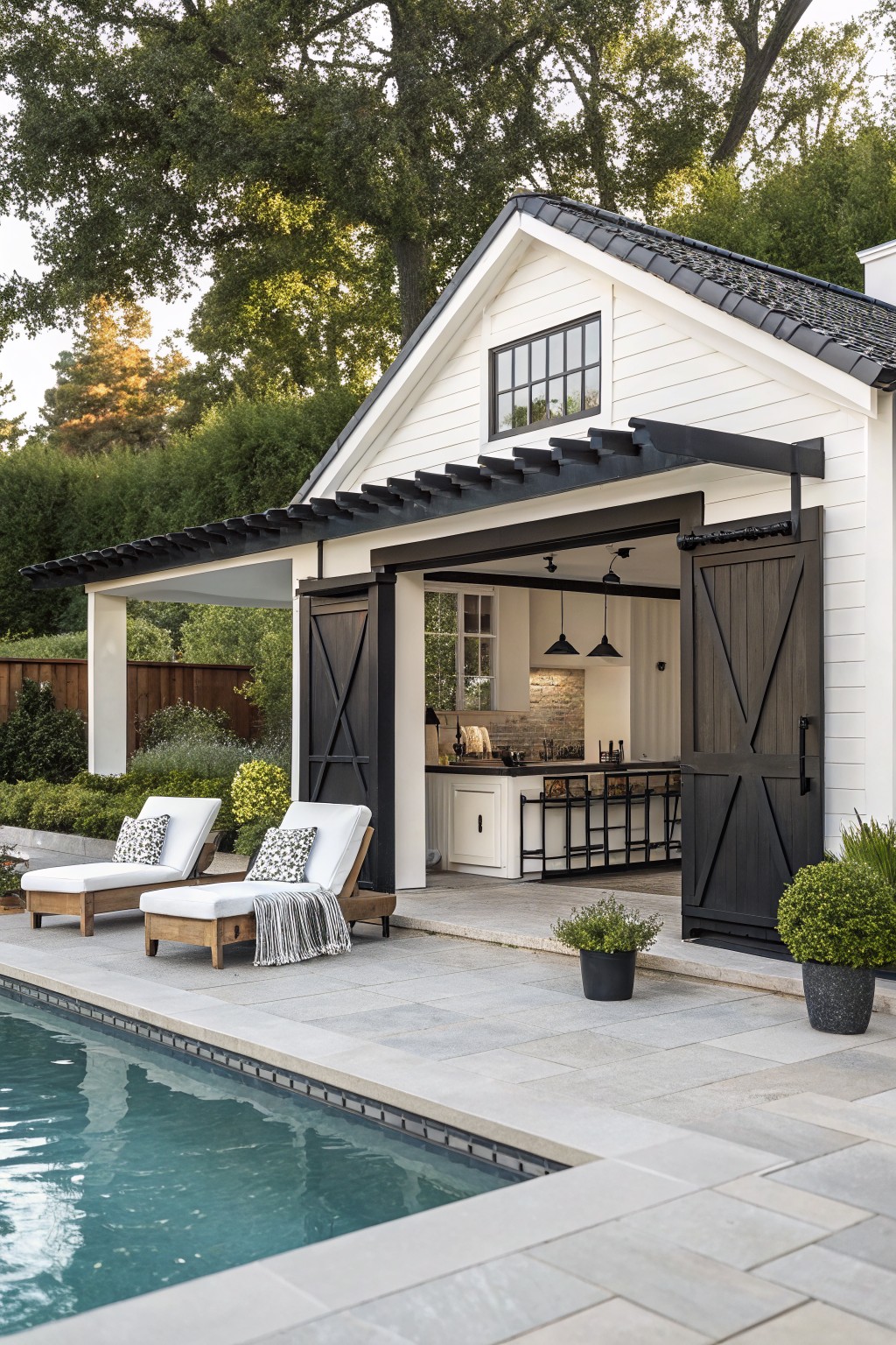 White pool house with open black barn doors revealing kitchen, lounge chairs by pool