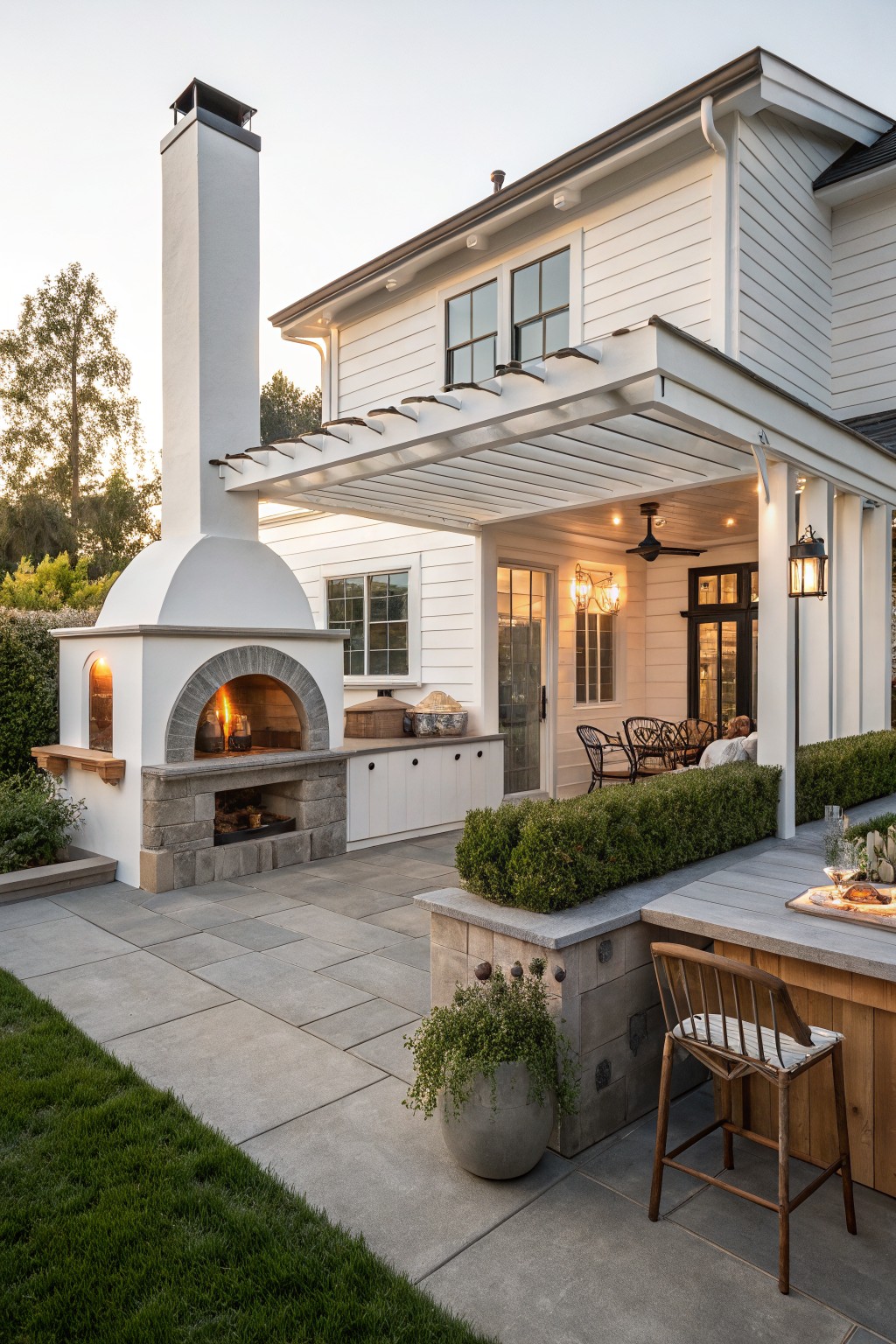 A white farmhouse with a large attached patio featuring a tall white outdoor fireplace and oven, a covered seating area, and a wooden bar with stools.