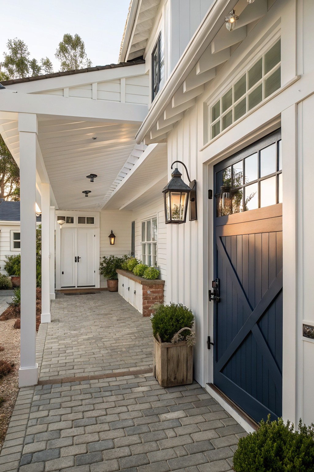 A white farmhouse exterior with a dark blue barn-style door, covered porch, and brick-paved walkway leading to double doors at the far end.