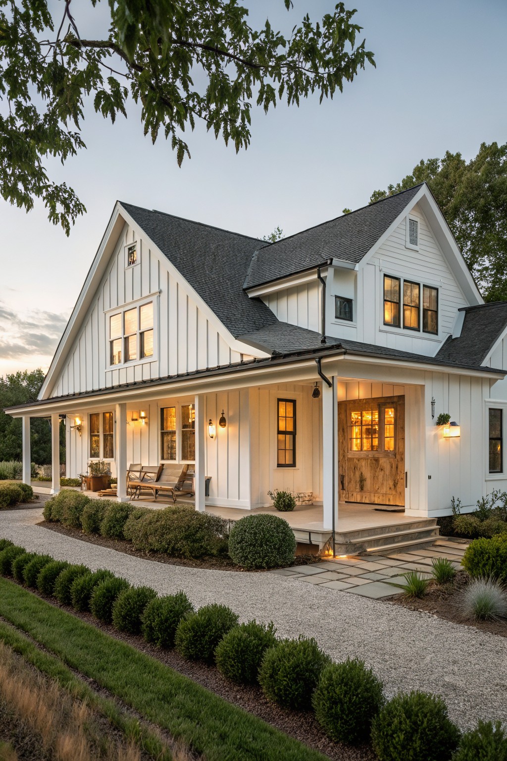 A white farmhouse ranch home with dark roofing, a covered front porch, and a wooden entry door illuminated at dusk.
