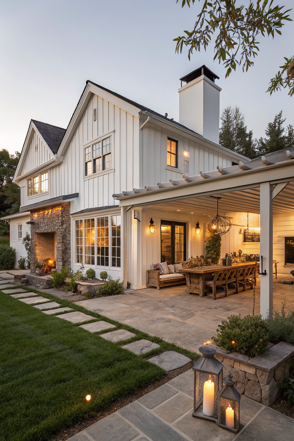 White farmhouse with stone chimney, covered patio, wooden table, and glowing lanterns.