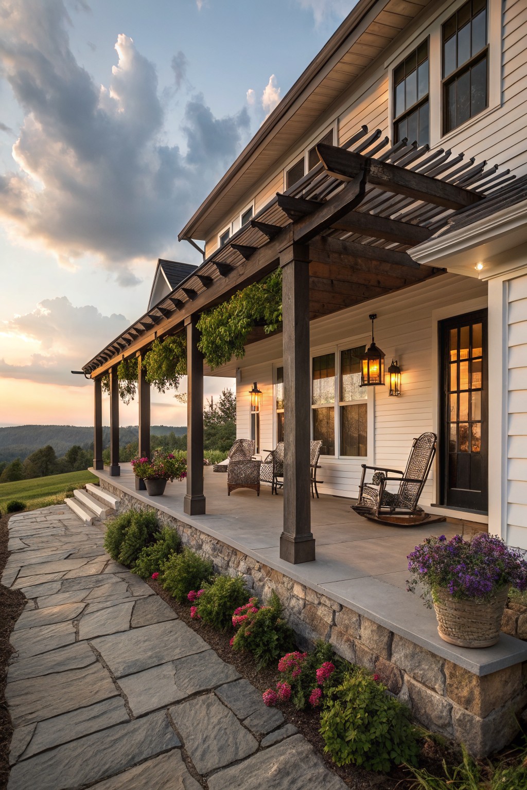 A white farmhouse exterior with a long covered porch supported by dark wood posts and a pergola, furnished with wicker rocking chairs and surrounded by potted flowers and stone landscaping.