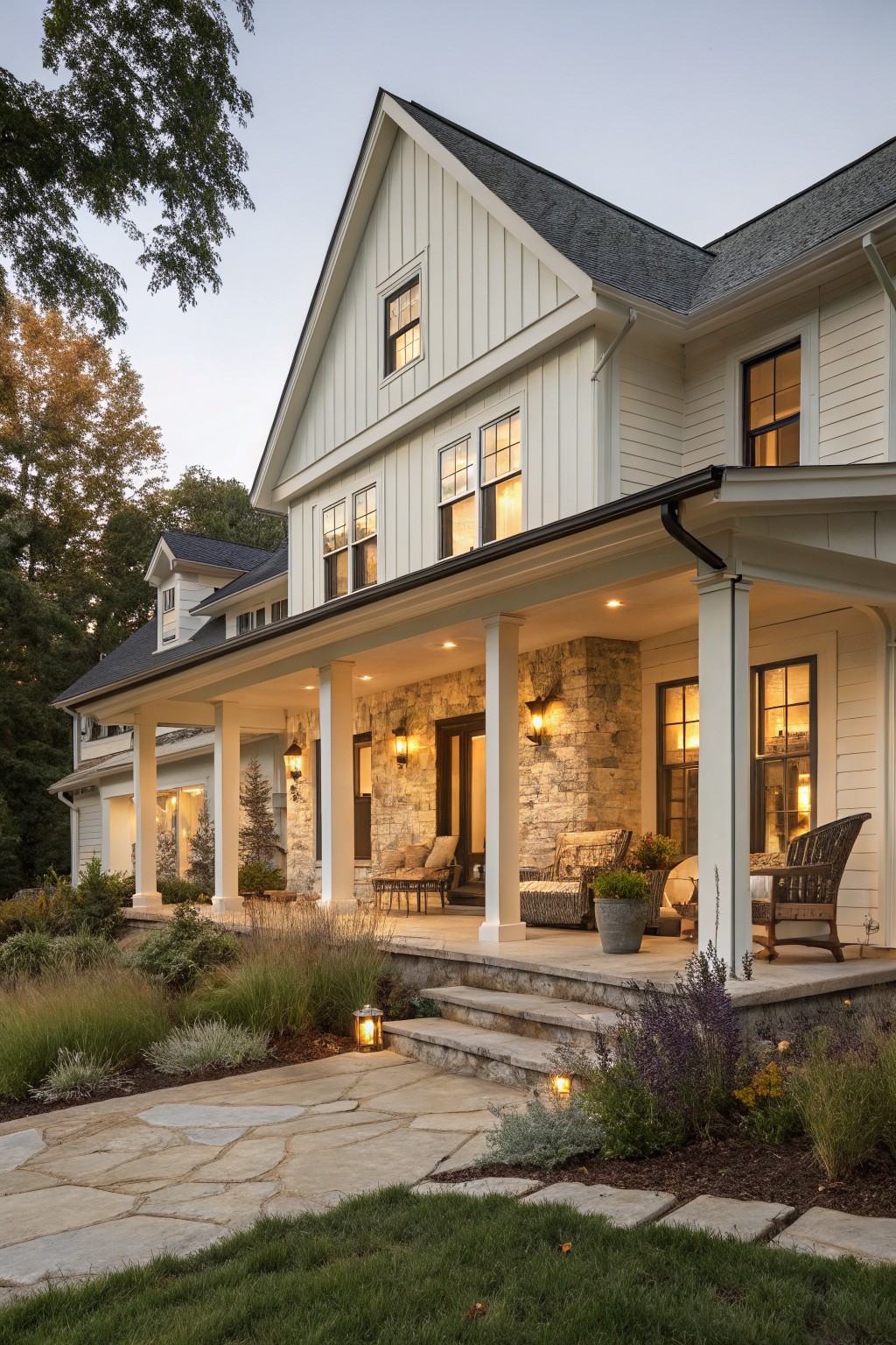 A two-story white farmhouse exterior at dusk with a wide covered porch, stone accent wall, white columns, and stone pathway in the foreground.