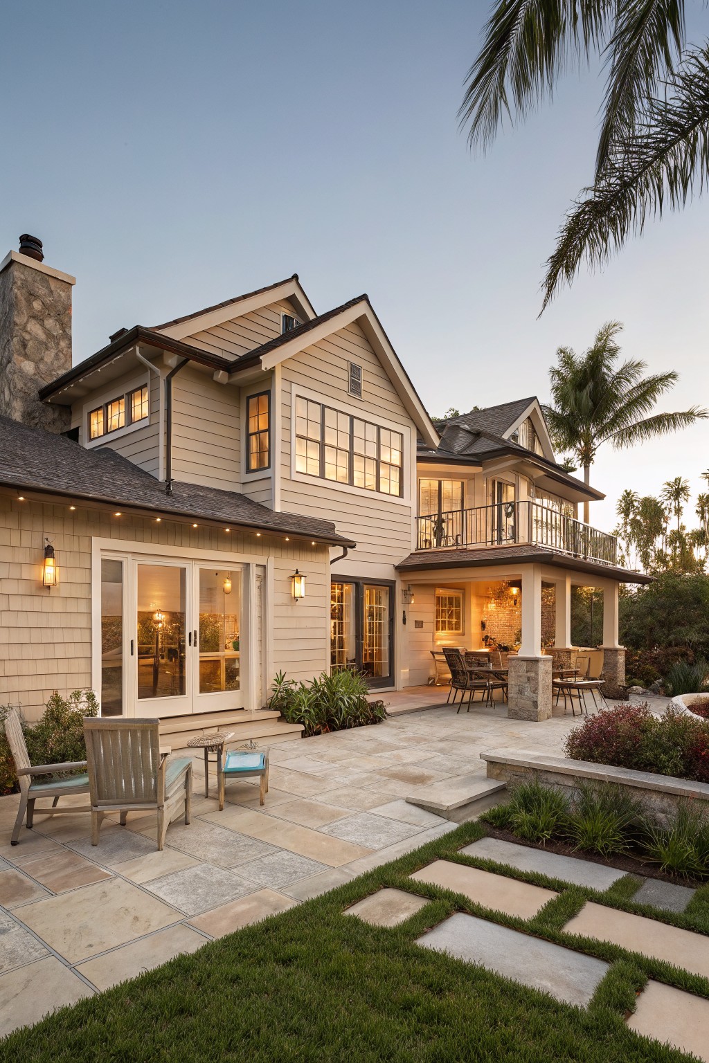 A two-story house with light shingle siding, white trim, large windows, and a stone patio with outdoor furniture.