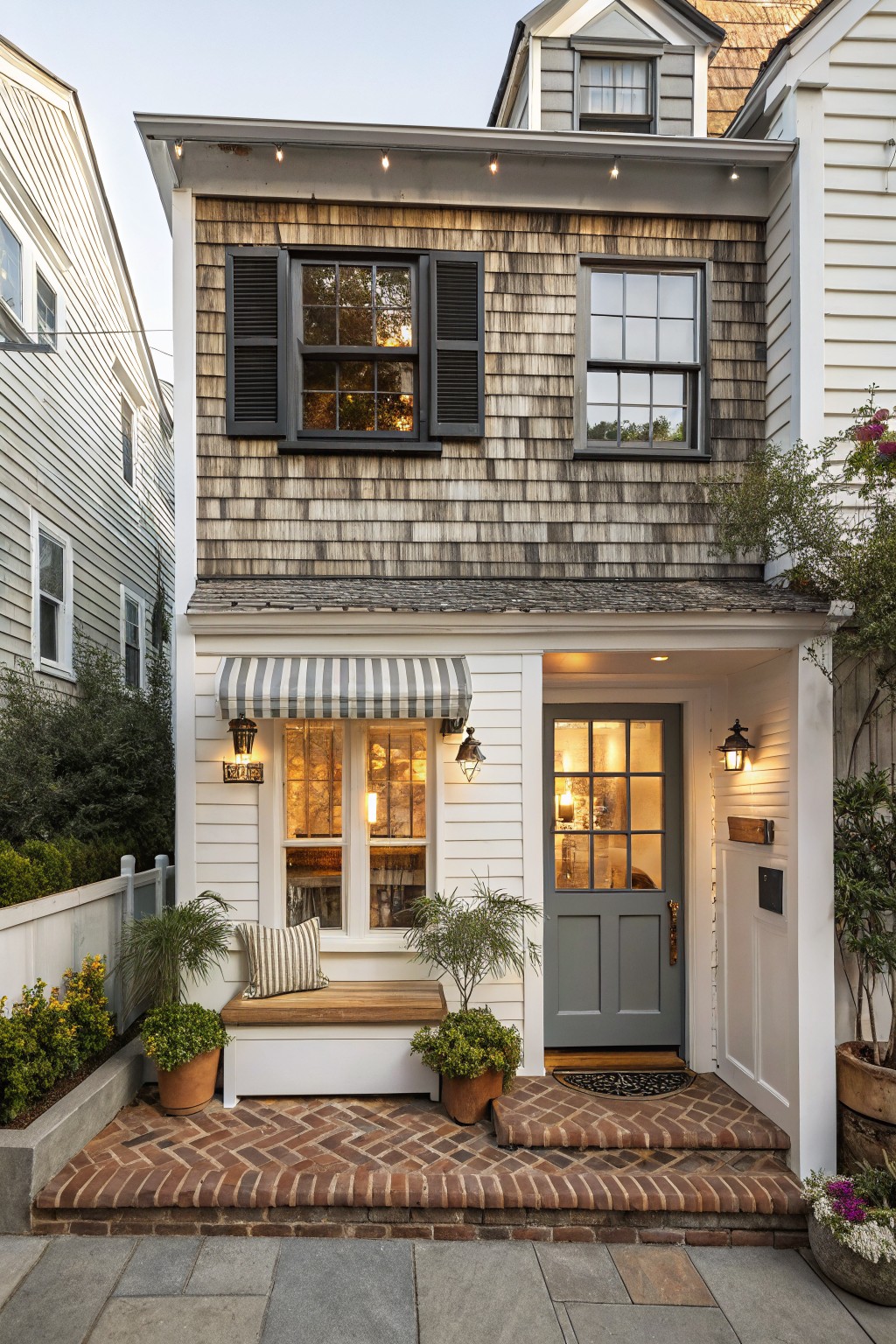 A two-story coastal house with weathered wood shingles on the upper facade and white painted siding below, featuring a gray front door, striped awning, brick steps, and black shutters on the windows.