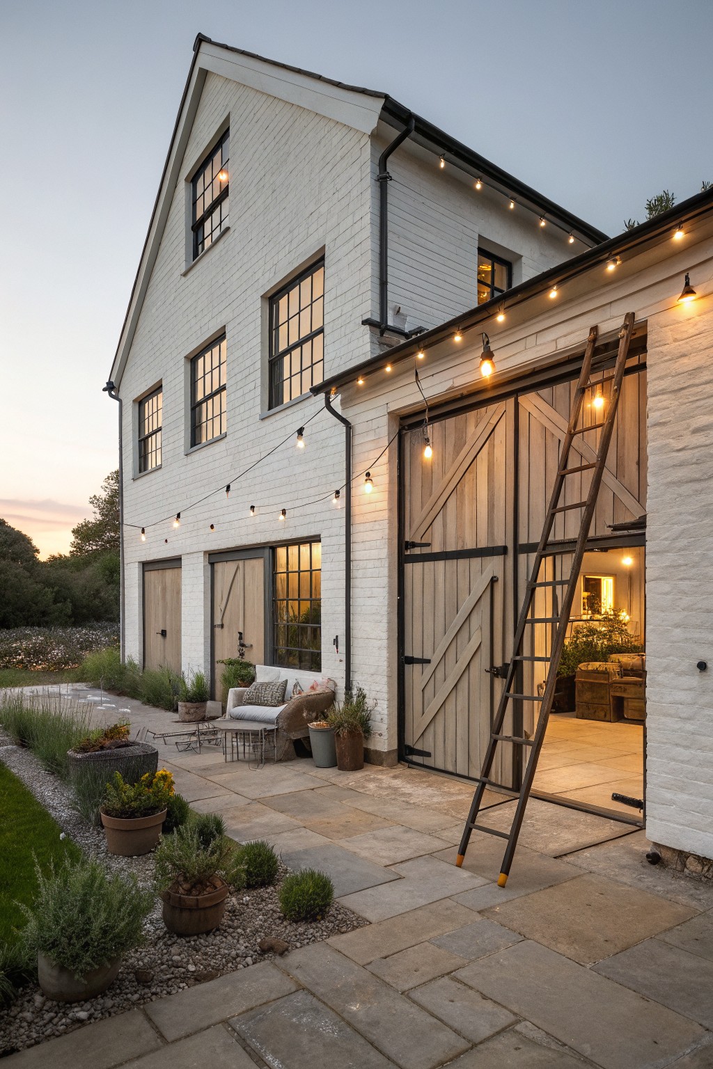 A two-story white farmhouse with black-trimmed windows, wooden barn doors, string lights along the eaves, and a stone patio with potted plants.