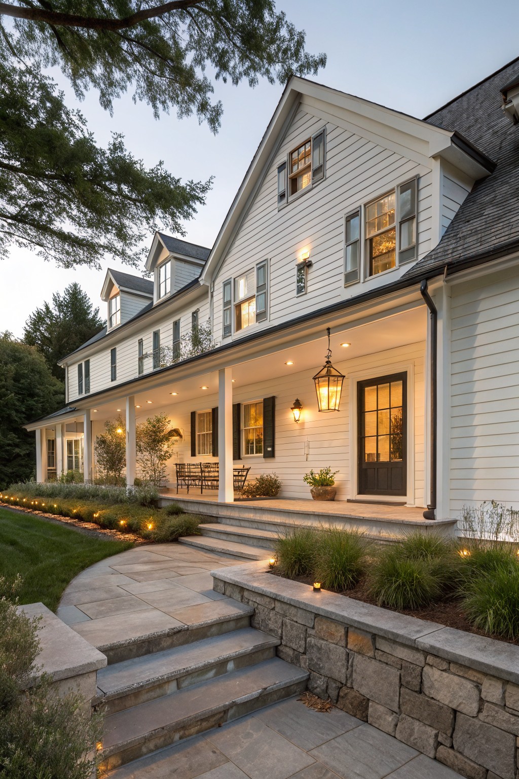 A two-story white farmhouse with a wide covered porch, dark window trim, stone steps, and warm exterior lighting along the path and beds at dusk.