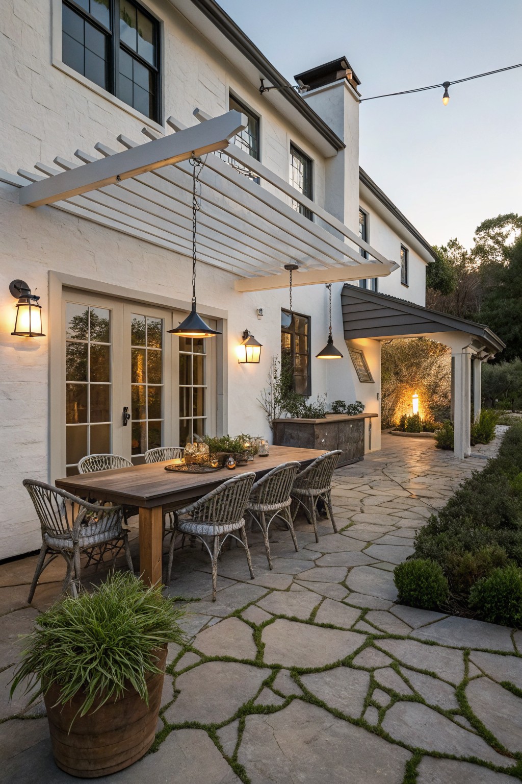 A creamy white farmhouse exterior featuring a wooden dining table and chairs arranged on a stone patio beneath a white pergola.
