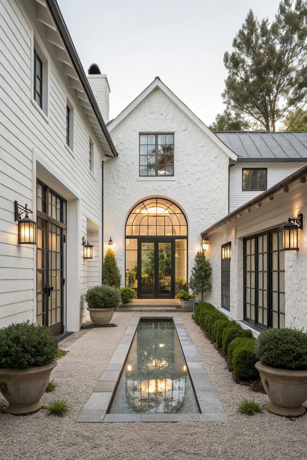 Front view of a white farmhouse with a large arched entryway of black-framed glass doors, flanked by windows and potted plants, with a long reflecting pool in the gravel courtyard.